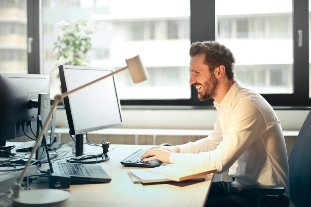 man in white dress shirt sitting on black rolling chair while facing black computer set and smiling